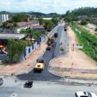 Imagem aérea mostra o início da pavimentação em estrada de chão. Ao lado da rua, há uma escola e do outro lado, um pequeno rio