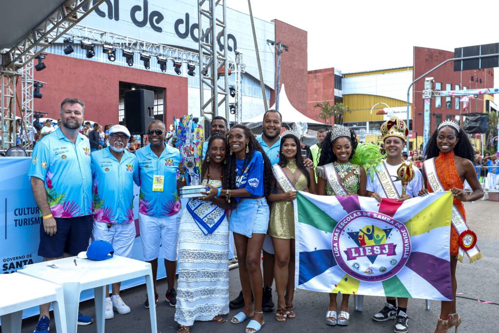 Duas mulheres negras sorriem e seguram um troféu. Ao redor, homens e mulheres também em pé sorriem