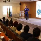 Homem com camisa azul está em pé, no palco de um auditório, segurando o microfone. Pessoas estão sentadas assistindo à apresentação