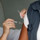 Mulher de óculos manuseia seringa com agulha e aplica vacina no braço de uma pessoa que usa colete de agente de endemias