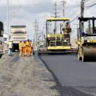 Caminhões, máquinas e homens trabalham em avenida que recebe novo asfalto
