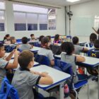 Sala de aula com professora a frente e alunos sentados em cadeiras na frente de carteiras