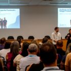 Homem e mulher estão em pé, a frente de um palco em que dois telões trazem informações sobre avaliação de desempenho. De costas para a câmera, há dezenas de homens e mulheres sentados assistindo à apresentação