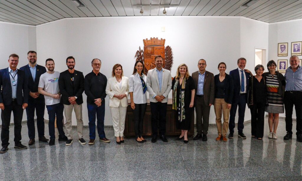 Homens e mulheres estão lado a lado, em pé, à frente do brasão em madeira da Prefeitura, e sorriem para foto