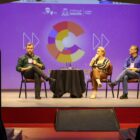 Em um palco, há uma mesa redonda no centro, com um homem sentado à esquerda e um homem e uma mulher sentados à direita