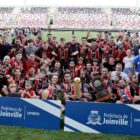 Várias pessoas estão uniformizadas em um estádio de futebol. Dois homens estão à frente e seguram um troféu cada