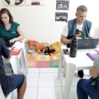 Duas pessoas sentadas em cadeiras na frente de computadores atendem outras duas pessoas também sentadas em cadeiras