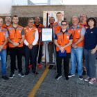 Homens e mulheres vestidos com coletes laranjas estão em pé, próximos à placa de metal