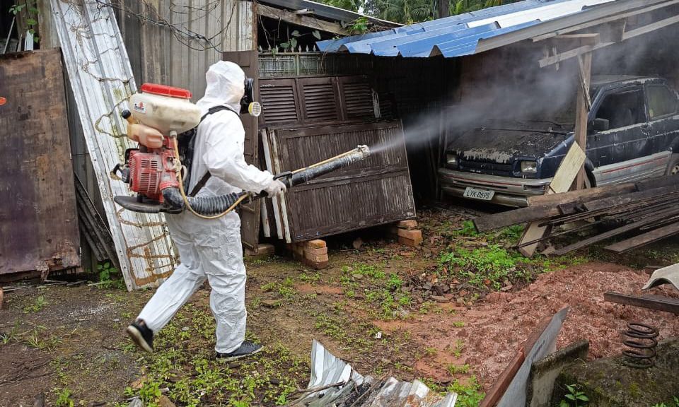Pessoa completamente vestida com equipamento de proteção branco leva pulverizador nas costas e aplica inseticida contra o mosquito da dengue em uma área com madeiras e telhas dispersas, além de um carro antigo
