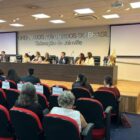 Evento com mesa diretiva com homens e mulheres, mulher discursa em púlpito e plateia observa sentada em cadeiras