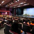 Duas mulheres estão no palco de um teatro enquanto dezenas de mulheres estão sentadas em poltronas de costas para a câmera