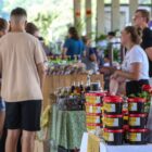 Em primeiro plano, há vários potes de doce de fruta dispostos de uma mesa, enquanto ao fundo pessoas conversam e sorriem