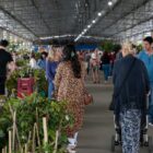 Diversas pessoas caminham por um grande galpão onde há exposição de plantas e outros produtos