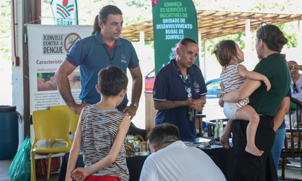 Um homem e uma mulher com uniforme da Vigilância Ambiental conversam com um homem, uma mulher segurando e um bebê e u menino
