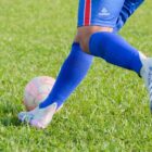 Imagem mostra as pernas de um homem com shorts e meia azul correndo em um gramado verde com uma bola próxima aos pés