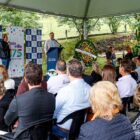 Homem fala em púlpito posicionado à frente, ao lado de uma coroa de flores, enquanto pessoas sentadas de costas para a câmera acompanham a sua fala