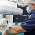 Homem de jaleco, máscara e gorro descartável manuseia equipamento médico enquanto paciente está deitado em uma máquina de exame