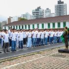 Centenas de meninos estão em fileiras lado a lado no pátio de um batalhão. Todos vestem camisas brancas e observam um homem à frente que veste uniforme militar e segura uma bandeira do Brasil