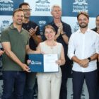 Homens e mulheres estão em pé, sorriam e posam para foto. Um homem e uma mulher seguram um documento.