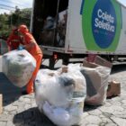 Dois homens vestidos com uniforme laranja colocam grandes sacos de lixo em um caminhão baú da coleta seletiva