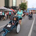 Em corrida de rua, mulher empurra cadeira de rodas com uma criança