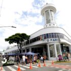 Imagem mostra imóvel pintado de branco com uma torre feita de concreto e vidro. Ao redor do prédio, há pessoas e tendas de uma feira