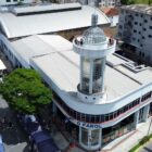 Imagem aérea mostra prédio branco na esquina entre duas ruas. O prédio tem uma torre construída em vidro e concreto e há pessoas no topo