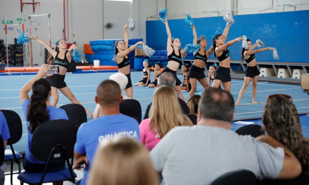 Em um espaço fechado, várias pessoas assistem a uma apresentação de ginástica artística feita por meninas