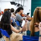 Imagem mostra mulher como maestra à frente e vários alunos, meninos e meninas, de costas para a câmera tocando instrumentos