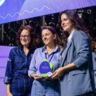 Três mulheres estão em um palco e sorriem para a câmera. Duas delas seguram um troféu