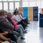 Homem branco está em pé, à frente de uma sala junto a um púlpito azul. Ele fala para uma plateia, cuja primeira fila aparece na foto, composta por homens e mulheres sentados