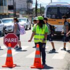 Alunos atravessam a faixa de pedestre enquanto outra aluna, vestida com colete e boné reflexivos, segura uma placa de Pare para interromper o trânsito de veículos
