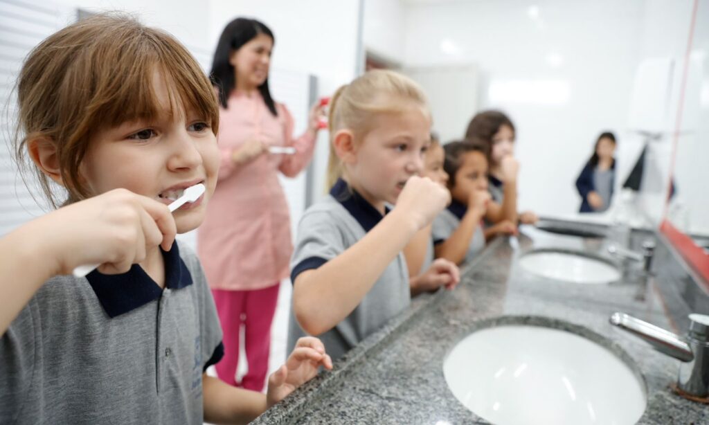 Quatro meninas escovam os dentes à frente de uma pia. Elas são supervisionadas por uma mulher vestida de rosa