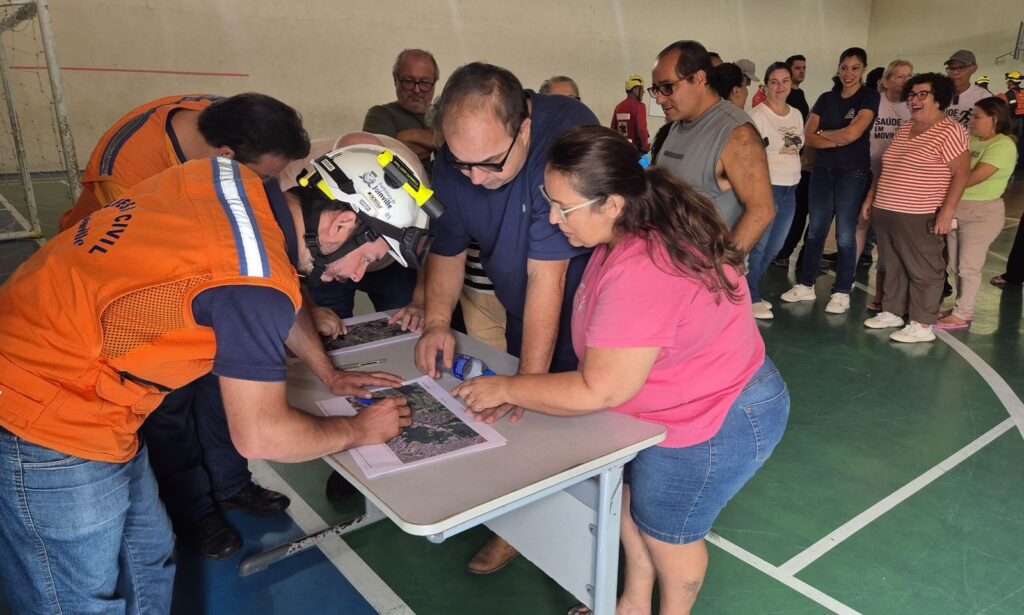 Homem com colete laranja e capacete olha para mapa junto a um homem e uma mulher