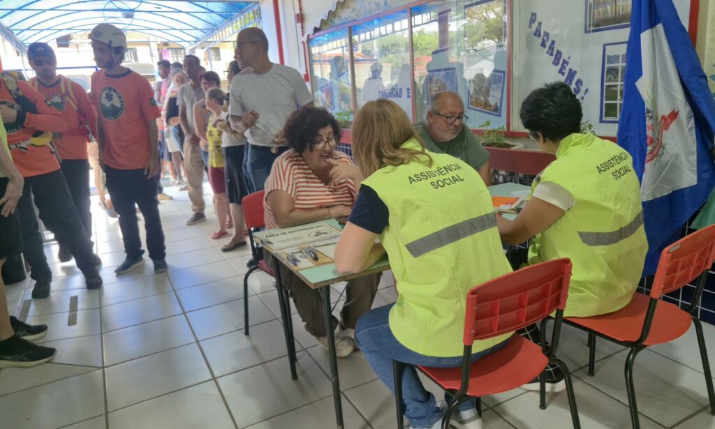 Duas mulheres de colete verde estão sentadas e atendem um homem e uma mulher sentados com uma fila de pessoas ao fundo