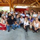 Dezenas de pessoas estão posando para a foto em um galpão. Algumas delas seguram um documento em papel branco