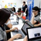 Em sala de aula, alunos adolescentes estão em computadores auxiliados por uma professora em pé