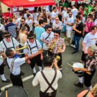 Pessoas vestidas com trajes típicos da Alemanha tocam instrumentos de sopro em meio à confraternização em uma rua repleta de pessoas