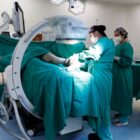 Imagem mostra um centro cirúrgico com equipamento em formato de arco em C. Há um paciente na mesa cirúrgica, mas apenas uma parte de sua perna está à mostra, enquanto um médico avalia o membro e outra pessoa o acompanha