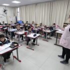 Professora em pé em sala de aula, fala com alunos sentados em cadeiras na frente de carteiras