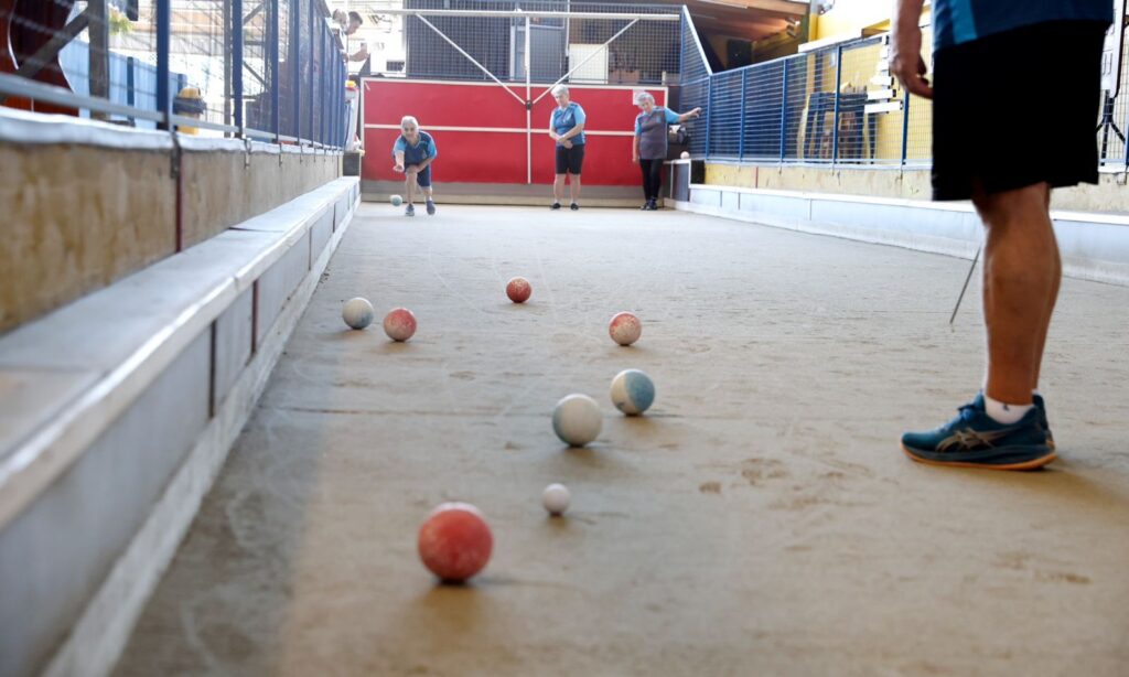 Imagem mostra uma quadra de bocha, com bolas de vários tamanhos posicionadas em primeiro plano e jogadores ao fundo