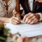 Imagem mostra parcialmente um homem e uma mulher vestidos de noivos. A mulher está assinando um documento