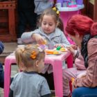 Mulher com cabelos vermelhos está sentada próxima a uma mesa infantil cor de rosa, onde três meninas estão brincando