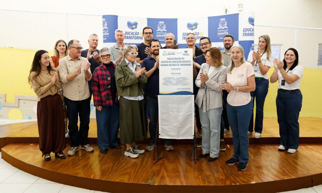 Homens e mulheres estão em pé enquanto aplaudem e sorriem. No centro, há uma placa que simboliza a entrega do CEI