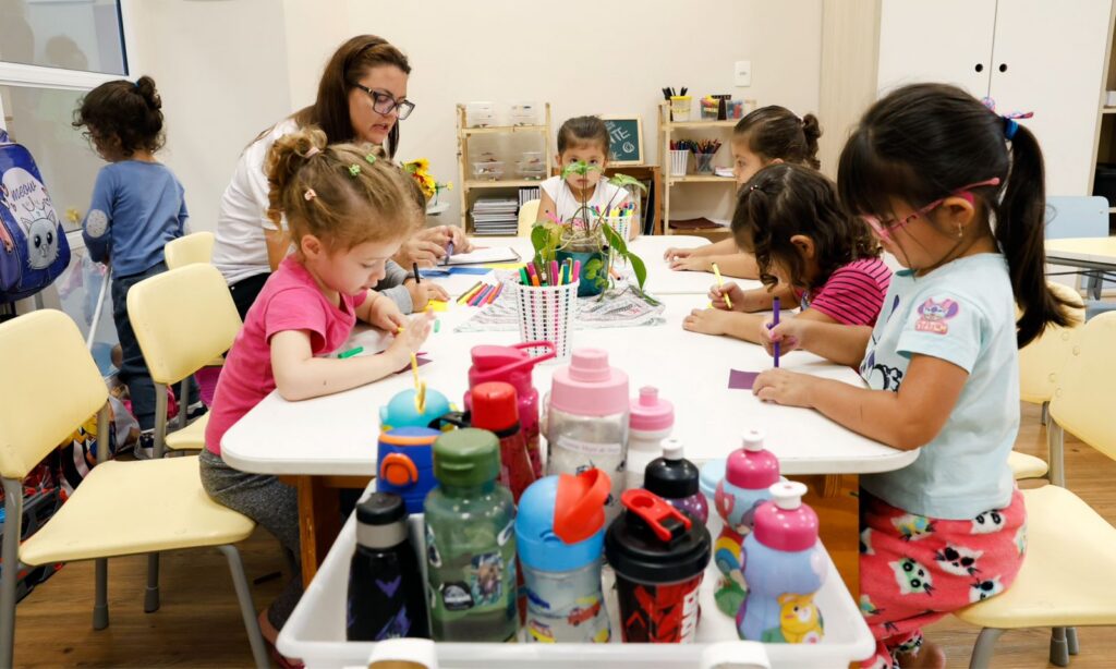 Em uma mesa branca, uma mulher acompanha várias meninas de cerca de três anos que desenham