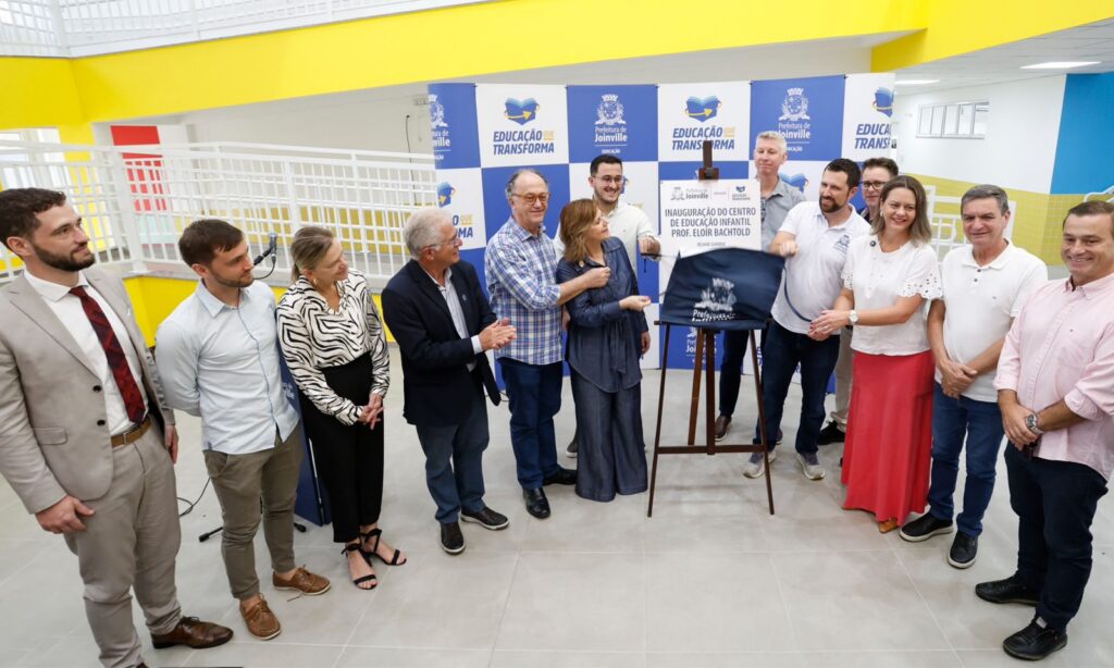 Homens e mulheres estão em pé, lado a lado, próximos a uma placa de metal com a escrita CEI Eloir Bachtold