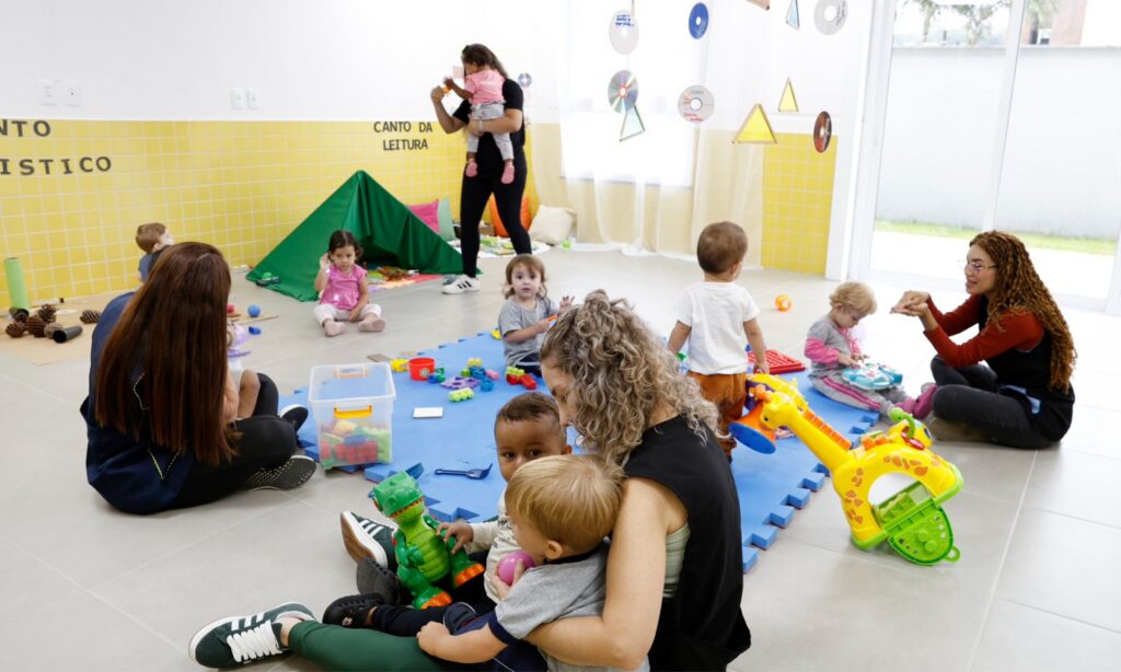 Em um espaço amplo, mulheres professoras brincam com bebês. Há barraca, pelúcias e outros brinquedos espalhados pela sala