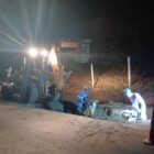 Imagem mostra homens trabalhando próximos a uma retroescavadeira em uma área aberta no solo durante a noite
