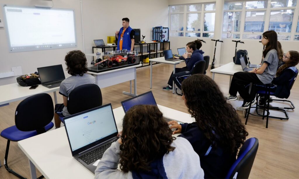Sala de aula com alunos consultando computadores com o professor à frente, em pé