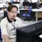 Jovem branco com cabelo curto está sentado à frente de computador e observa a tela